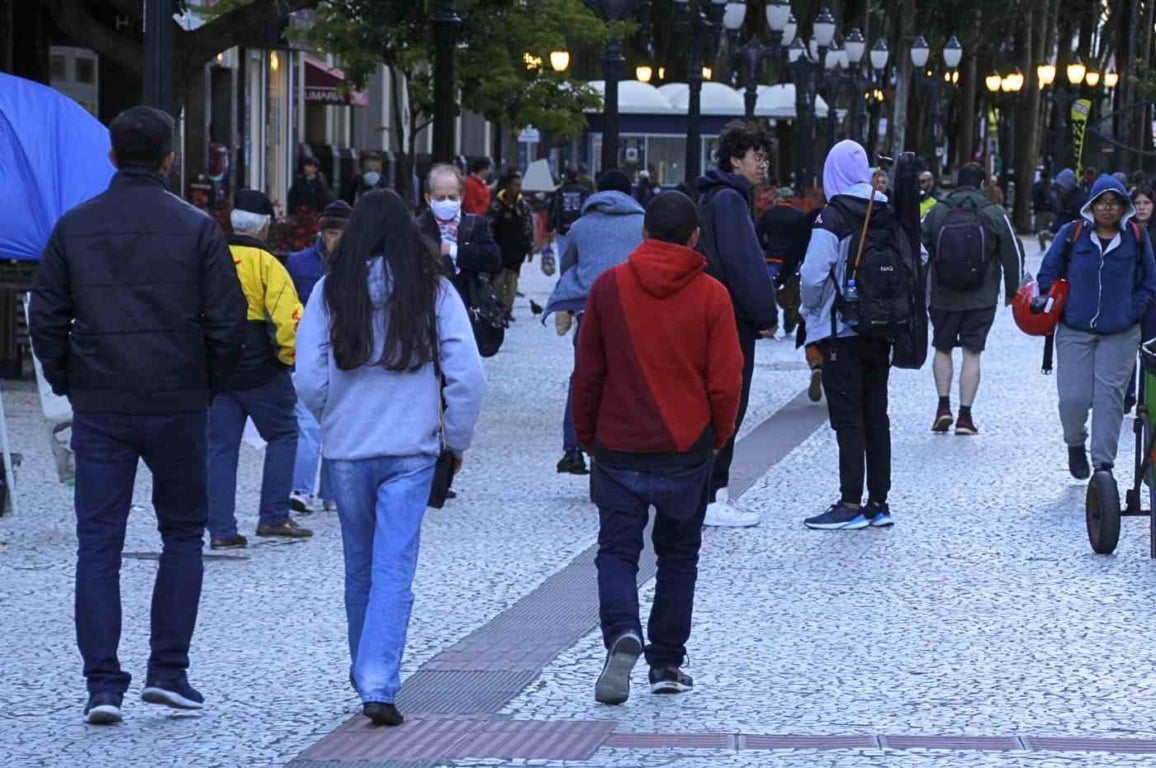 Pessoas caminham agasalhadas em calçadão urbano durante dia frio, com clima nublado e iluminação de postes acesa.