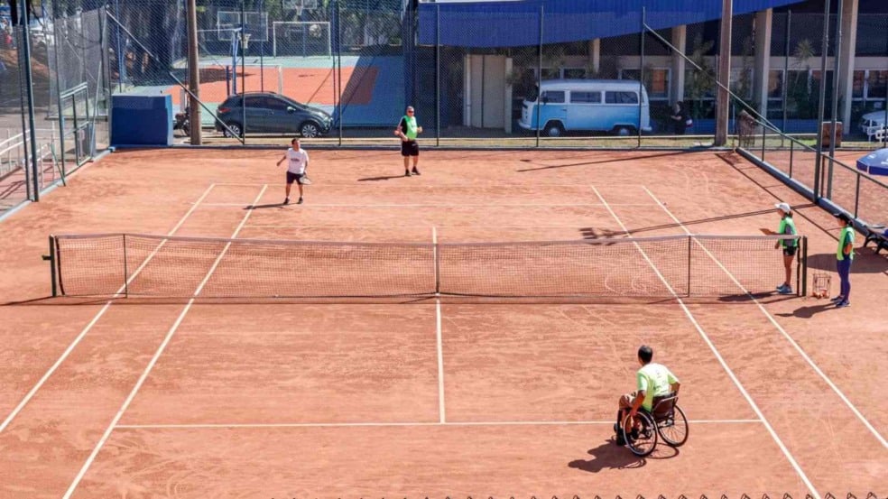 Jogadores de tênis, incluindo um atleta cadeirante, participam de partida em quadra de saibro no Festival Regional de Tênis em Jundiaí, acompanhados por instrutores na lateral.