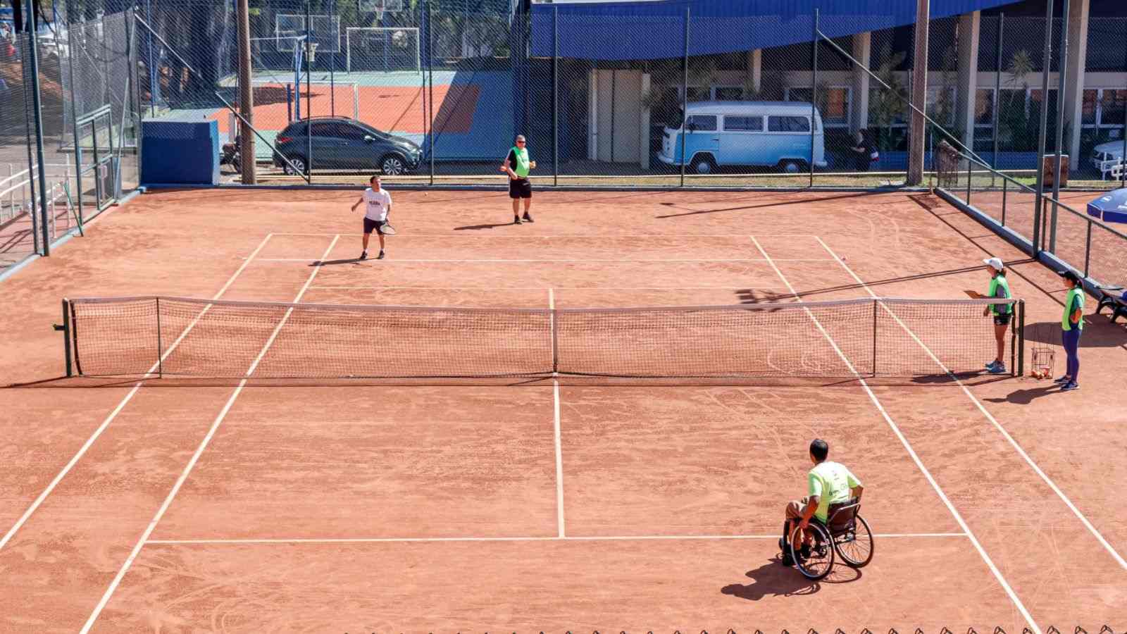 Jogadores de tênis, incluindo um atleta cadeirante, participam de partida em quadra de saibro no Festival Regional de Tênis em Jundiaí, acompanhados por instrutores na lateral.