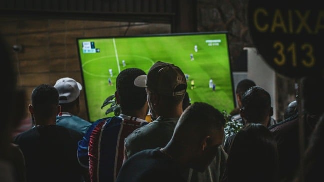 Grupo de amigos reunidos em frente a uma televisão assistindo a um jogo de futebol, mostrando o entusiasmo dos brasileiros por eventos e apostas esportivas.
