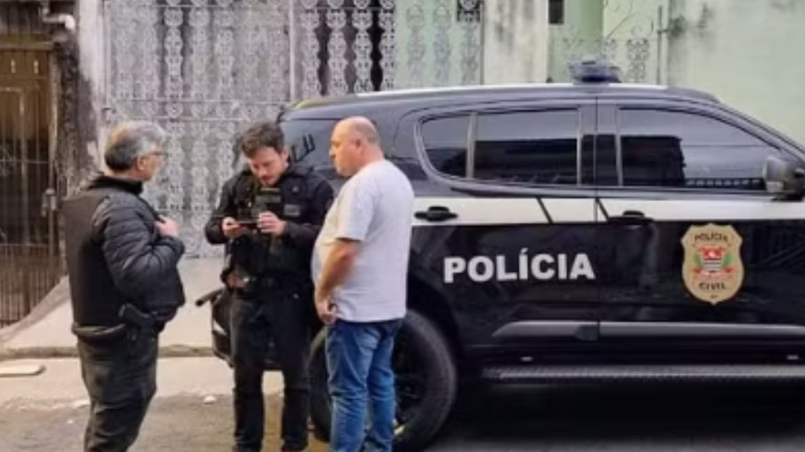 Policiais civis conversam com um homem ao lado de uma viatura preta da Polícia Civil em rua residencial.