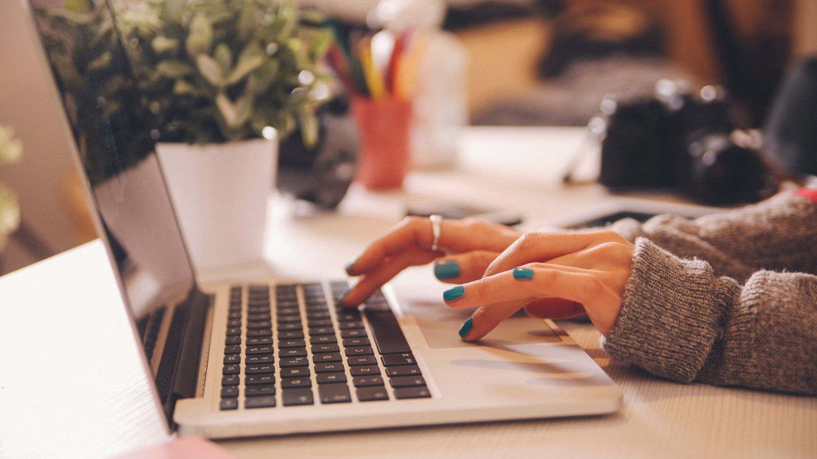 Mãos femininas digitando em um notebook sobre uma mesa de escritório com objetos coloridos ao fundo.