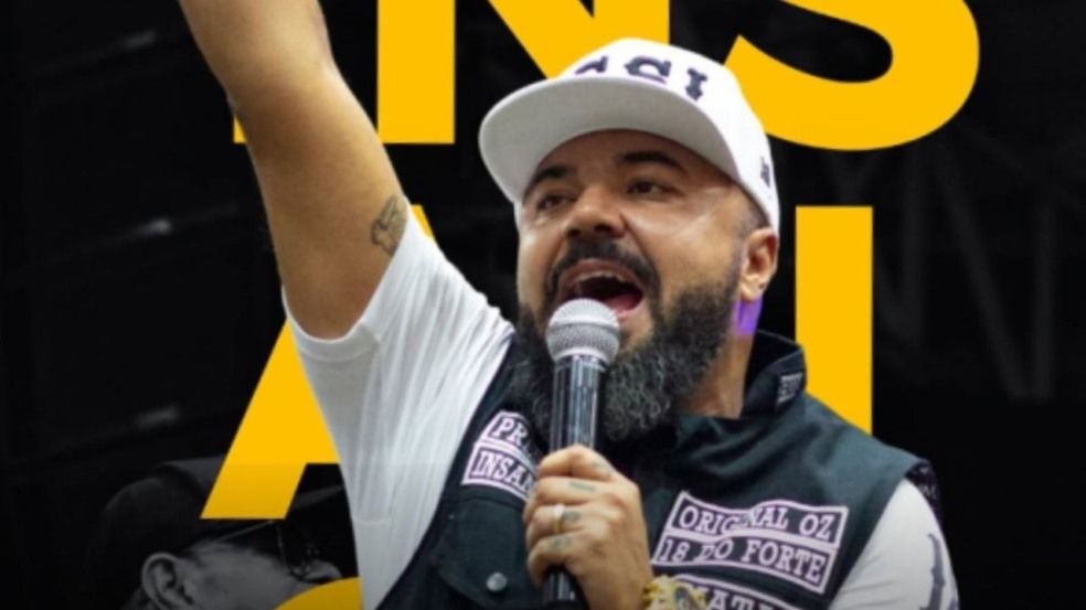 Homem com boné e microfone em evento, com letras amarelas de fundo.