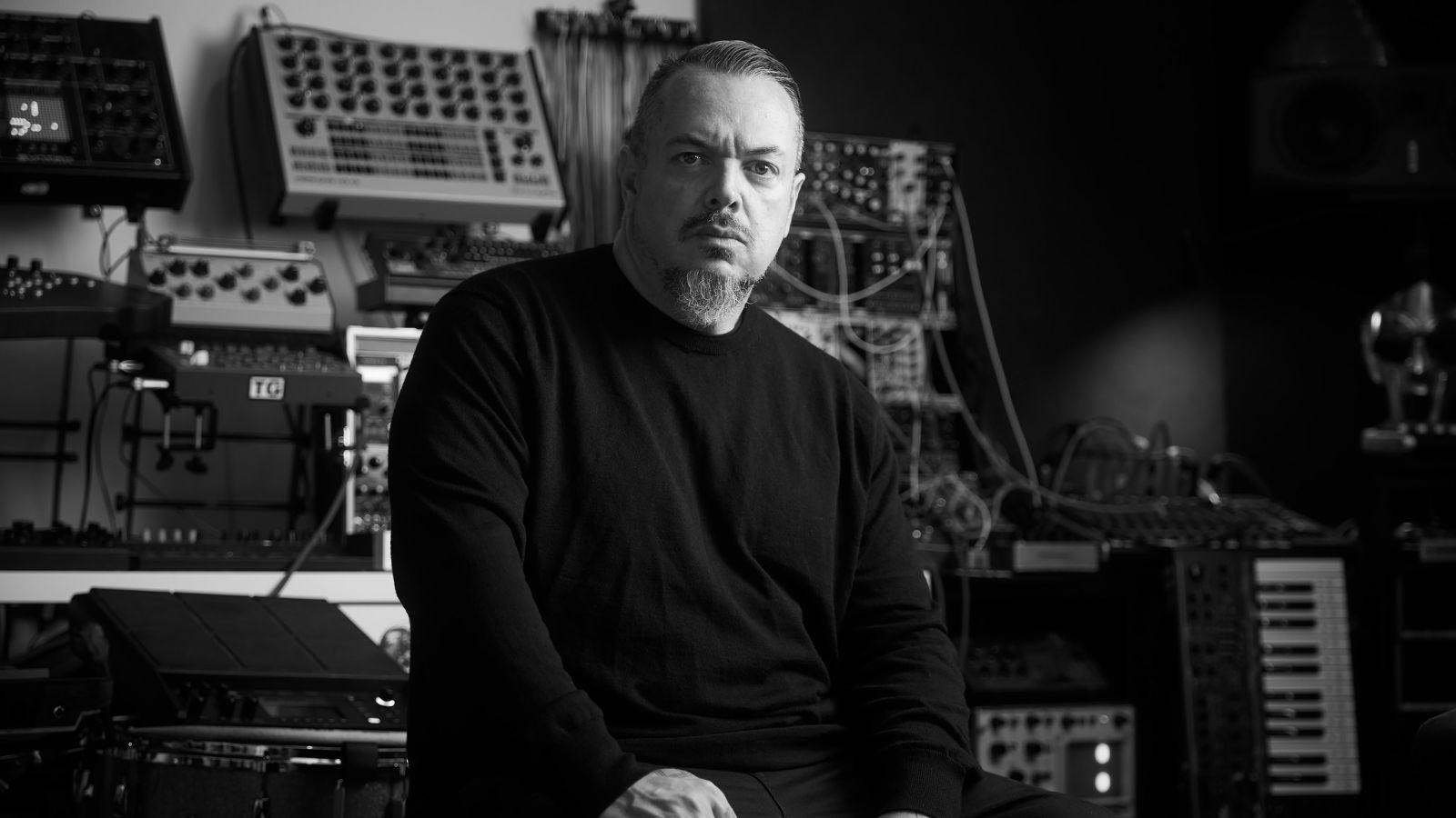 Iggor Cavalera sentado em estúdio, cercado por sintetizadores e equipamentos musicais, em foto em preto e branco.