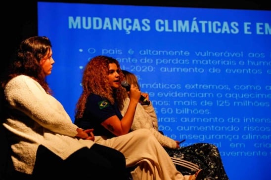 Mulheres discutem políticas para crianças e mudanças climáticas em painel, com apresentação em tela ao fundo destacando os riscos.