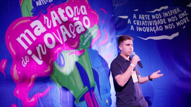 Jovem apresenta palestra na Maratona de Inovação em Jundiaí, diante de painel com frases sobre criatividade e sustentabilidade.