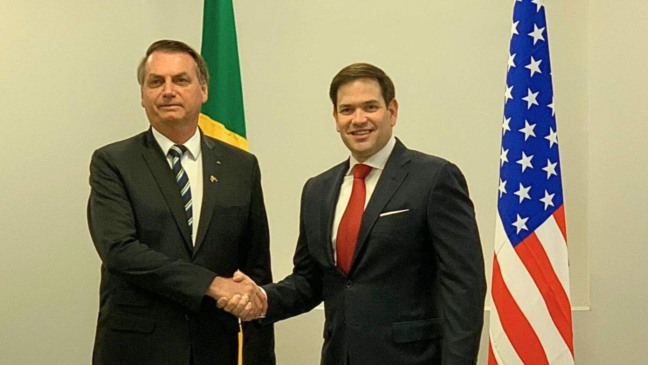 Jair Bolsonaro e Marco Rubio apertam as mãos em frente às bandeiras do Brasil e dos Estados Unidos.