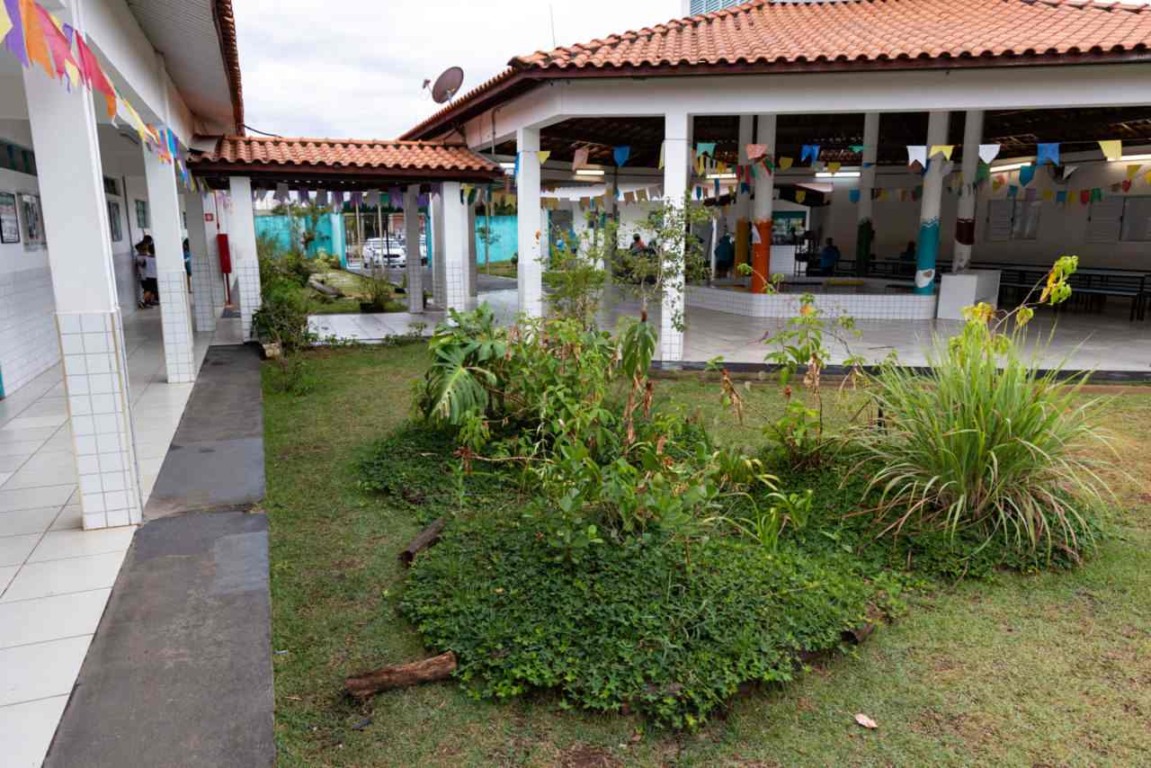 Vista do pátio de uma escola decorada com bandeirinhas coloridas, com um jardim central cercado por colunas e telhados de cerâmica.