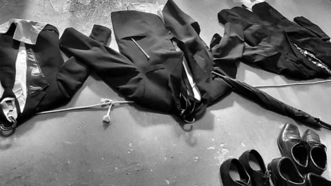 Roupas de cena em preto e branco dispostas no chão — paletós, camisas, sapatos sociais e guarda-chuva — representando elementos do espetáculo “João das Ruas, do Rio, da Gente Alegre”.