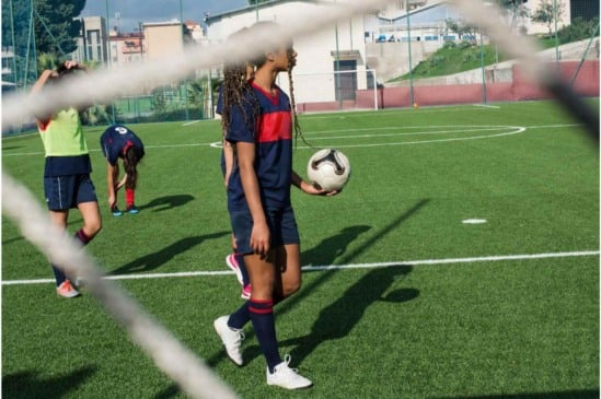 Jogadoras de futebol treinam em campo de grama sintética sob sol forte, com uma segurando a bola e outra ajustando o uniforme.