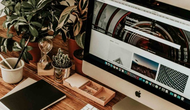 Espaço de trabalho organizado com iMac, plantas, caderno, ampulheta e acessórios, representando produtividade e design natural.