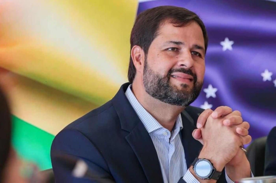 Luiz Fernando Machado sorrindo, de barba e terno azul, com as mãos unidas, frente a bandeiras desfocadas, uma com estrelas brancas.