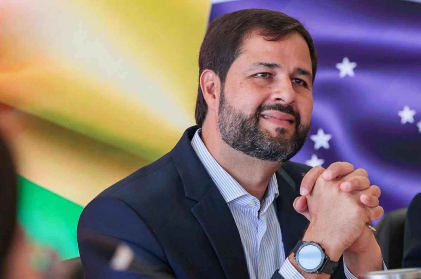 Luiz Fernando Machado sorrindo, de barba e terno azul, com as mãos unidas, frente a bandeiras desfocadas, uma com estrelas brancas.