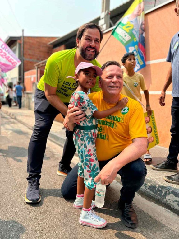 Parimoschi e Luiz Fernando abraçam uma criança durante campanha eleitoral no Jardim Tamoio, em Jundiaí, com apoiadores ao fundo.