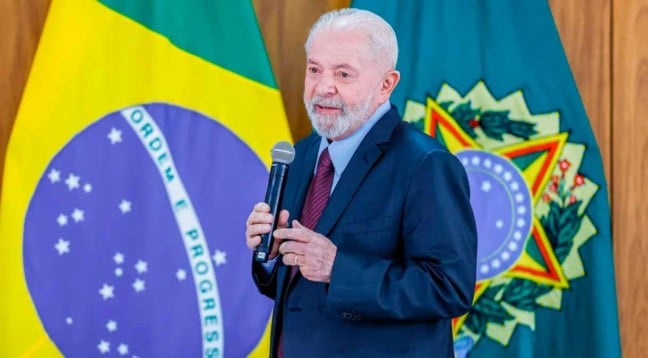 Presidente Lula falando ao microfone a frente da bandeira do Brasil