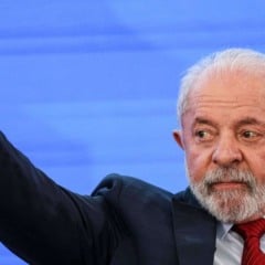 Presidente Luiz In&aacute;cio Lula da Silva, de terno e gravata vermelha, aponta para cima com o dedo indicador durante evento, em frente a um painel azul.