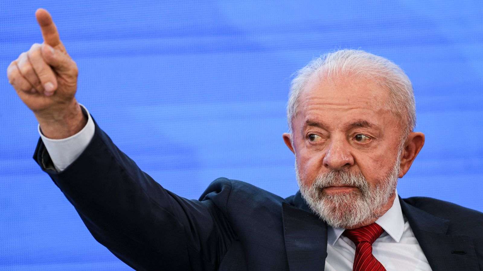 Presidente Luiz Inácio Lula da Silva, de terno e gravata vermelha, aponta para cima com o dedo indicador durante evento, em frente a um painel azul.
