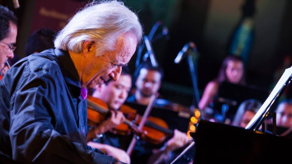 Maestro João Carlos Martins, se apresentando em um concerto, tocando piano. Ao fundo, membros da Bachiana Filarmônica estão tocando instrumentos de corda, como violinos. Maestro João Carlos Martins, se apresentando em um concerto, tocando piano. Ao fundo, membros da Bachiana Filarmônica estão tocando instrumentos de corda, como violinos.