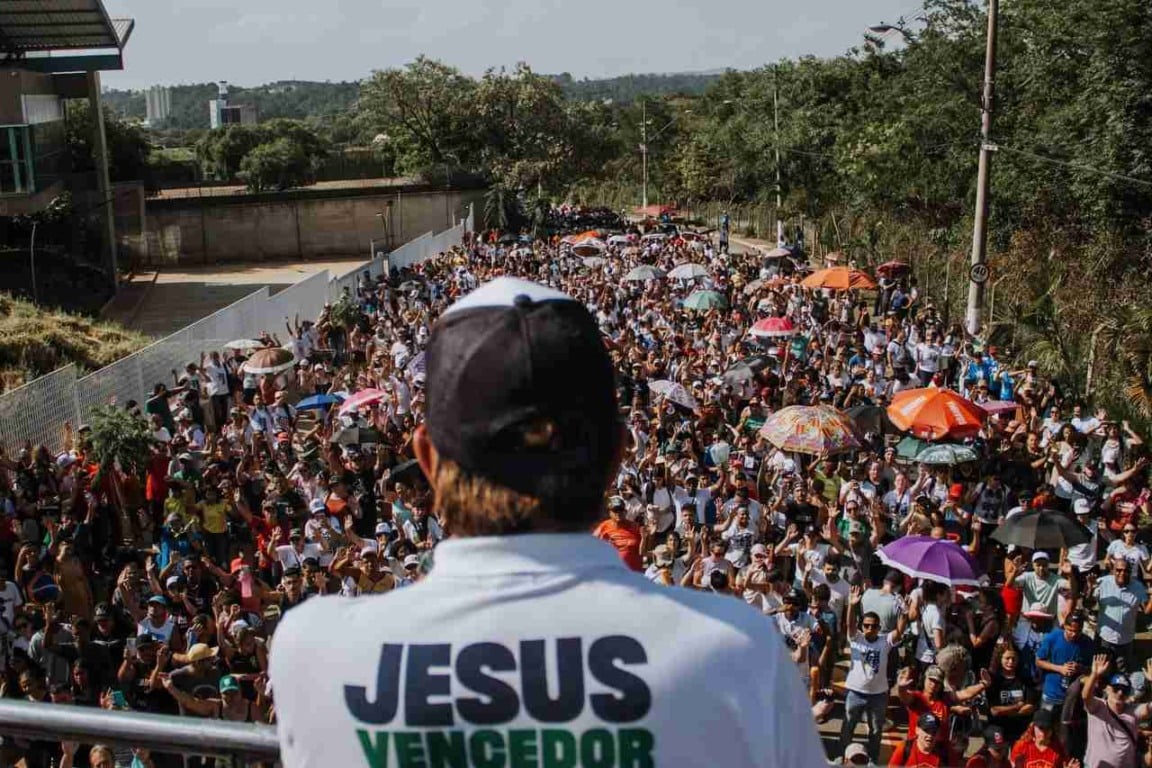 Líder de costas com camiseta "Jesus Vencedor" discursando para multidão na Marcha para Jesus, com pessoas e guarda-chuvas coloridos.