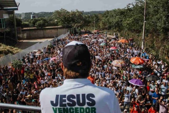 Líder de costas com camiseta "Jesus Vencedor" discursando para multidão na Marcha para Jesus, com pessoas e guarda-chuvas coloridos.