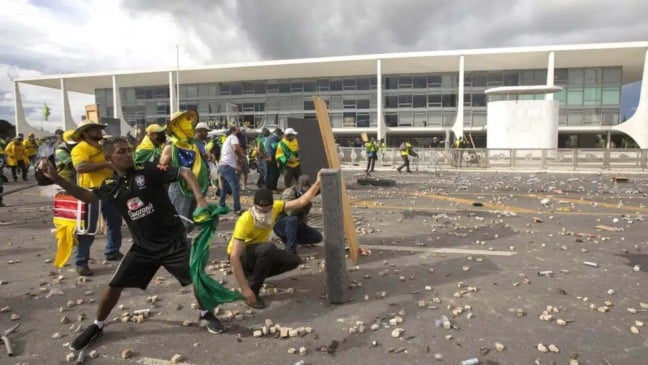 Manifestantes em frente à Praça dos Três Poderes, em Brasília, com alguns participantes jogando objetos e outros se protegendo com barricadas improvisadas nos atos golpistas de 8 de janeiro.