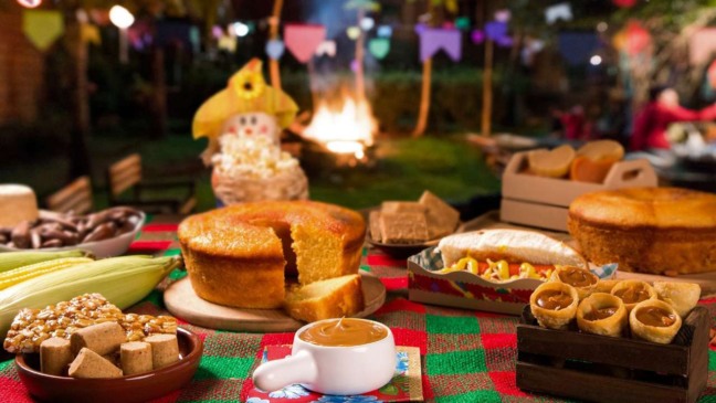 Mesa farta com comidas típicas de festa junina, como bolo de milho, paçoca, e canjica, com decoração festiva.