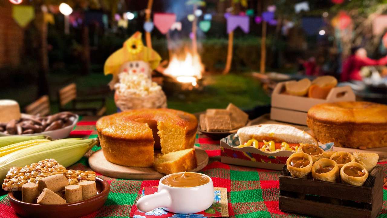 Mesa farta com comidas típicas de festa junina, como bolo de milho, paçoca, e canjica, com decoração festiva.