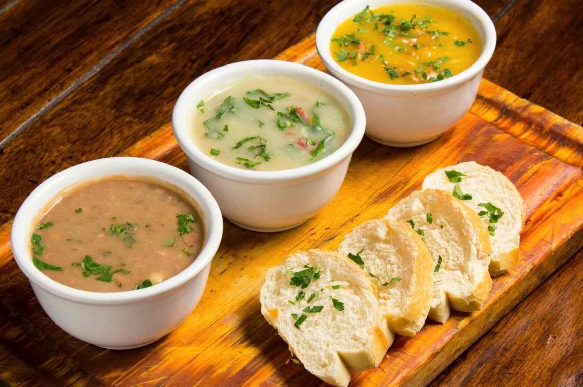 Três cumbucas de caldo de feijão, caldo verde e caldo de mandioquinha, acompanhadas de torradas