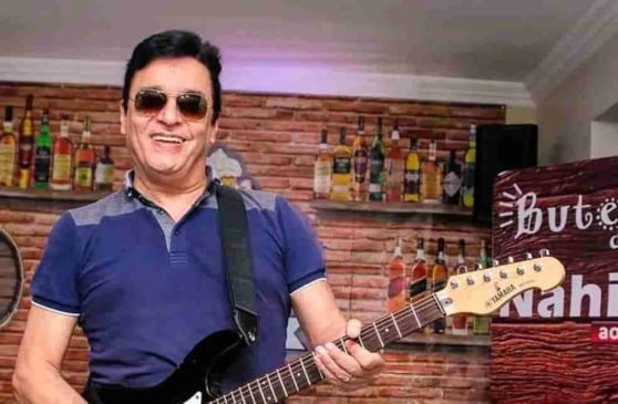 Cantor Nahin de camiseta azul segurando uma guitarra em um bar de fundo.