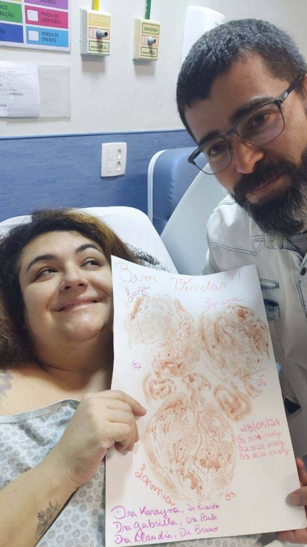 Pais sorrindo no hospital, segurando cartaz com impressões dos pés das filhas trigêmeas