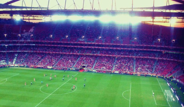Estádio de futebol iluminado ao entardecer, com jogadores em campo durante uma partida, enquanto o VAR é utilizado para revisões.