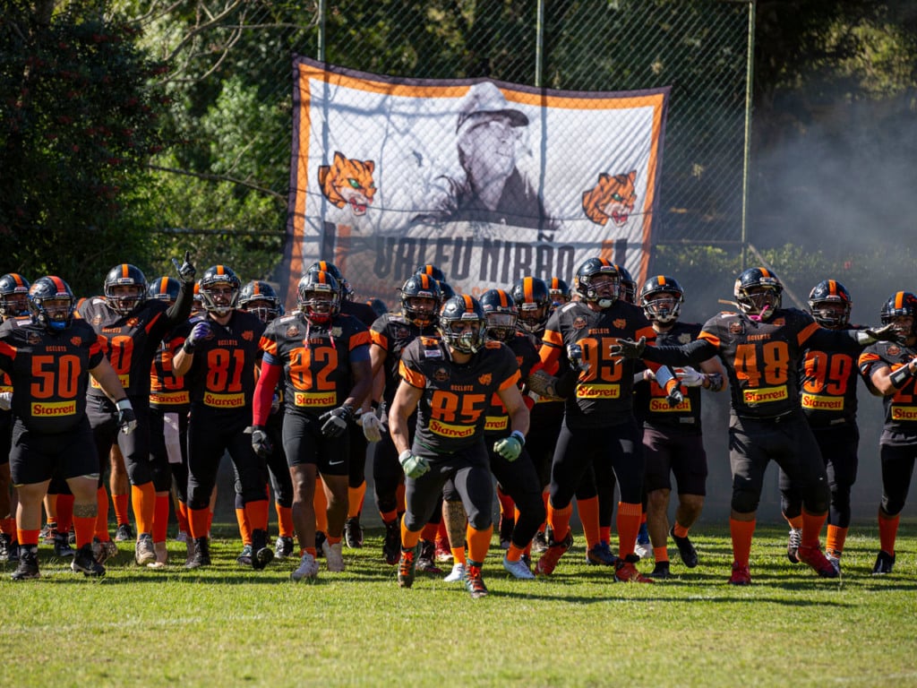 Equipe do Ocelots Futebol Americano entrando em campo com uniformes pretos e laranja antes do jogo, com bandeira do time ao fundo