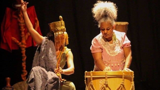 Atriz com adereço dourado e figurino brilhante ao lado de percussionista em traje rosa, durante apresentação do espetáculo Odus inspirado na mitologia iorubá.