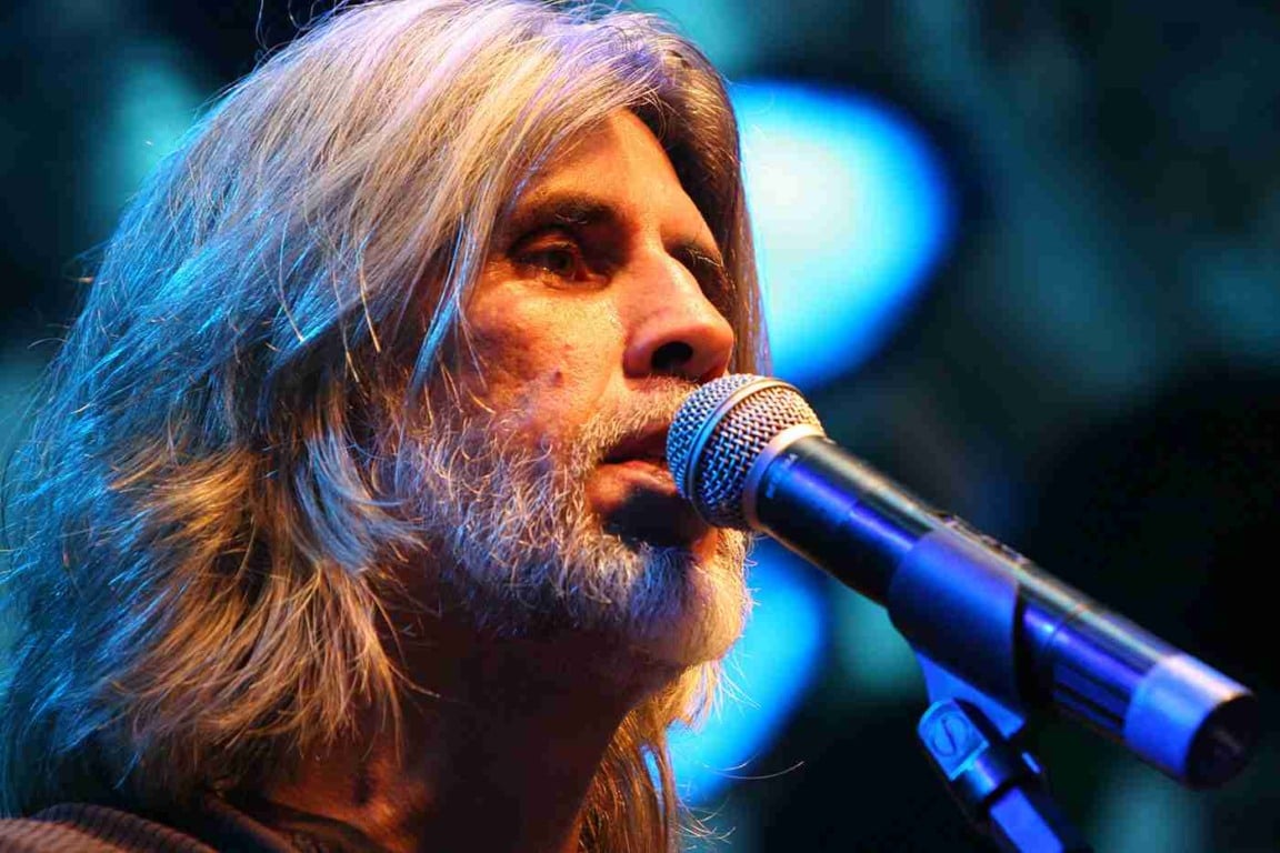 Osvaldo Montenegro cantando no microfone em um show, com cabelo e barba grisalhos, iluminação azul de fundo.