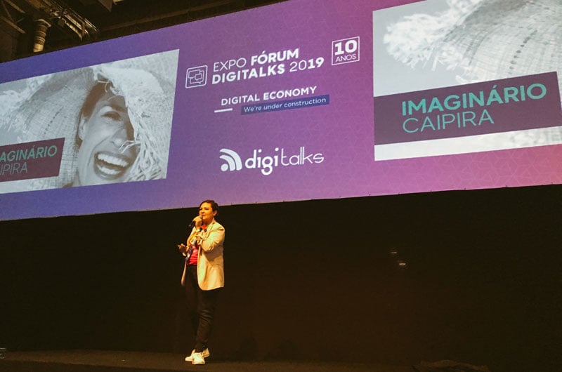Paola Faria palestrando no palco da Expo Fórum Digital 2019; ela está com blazer bege, calça preta, camiseta rosa e tênis branco, segurando microfone na frente de painel roxo.
