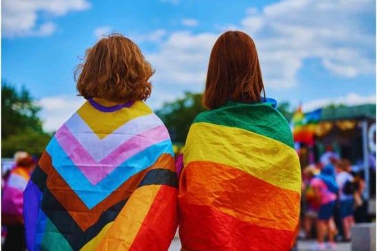 Duas pessoas de costas usando bandeiras do orgulho LGBTQIA+ na Parada LGBT+ 2024, uma com a bandeira do orgulho progressista e outra com a bandeira do arco-íris.