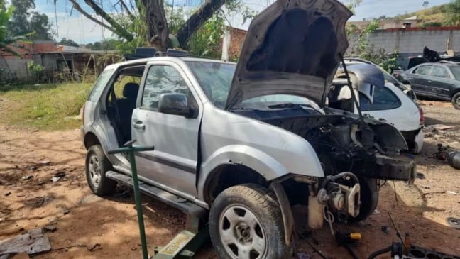 Veículo prateado parcialmente desmontado em um terreno aberto de desmanche clandestino em Campo Limpo Paulista. O capô do carro está levantado, expondo o motor, e as portas estão removidas.