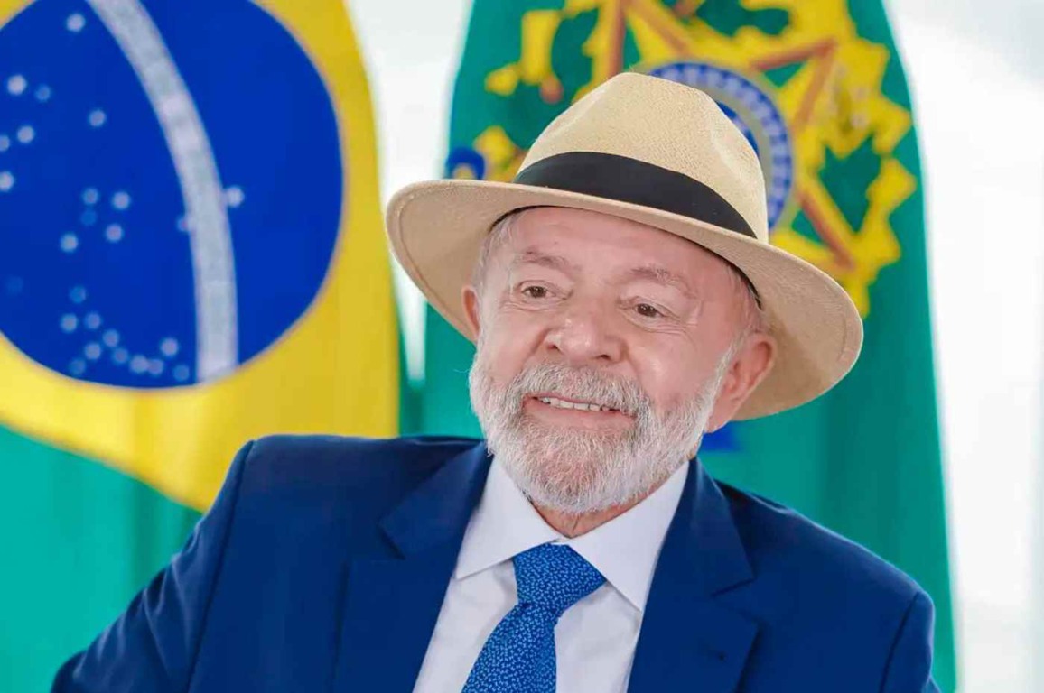 Presidente Lula sorrindo com chapéu à frente das bandeiras do Brasil