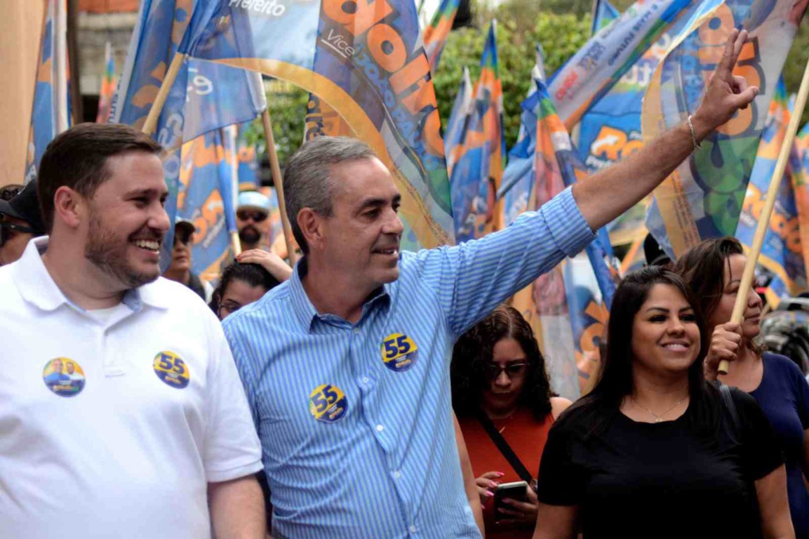 Professor Rodolfo, candidato à reeleição em Várzea Paulista, acena durante caminhada eleitoral ao lado de apoiadores, segurando bandeiras de campanha.