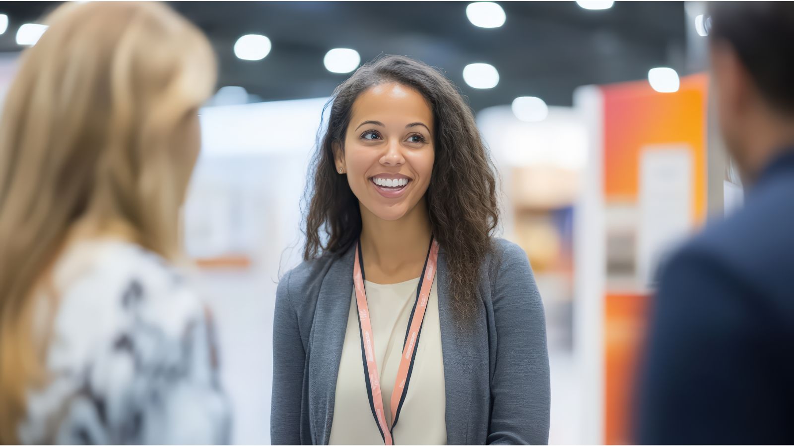 Mulher sorridente de blazer cinza com crachá conversando com duas pessoas em um evento corporativo.
