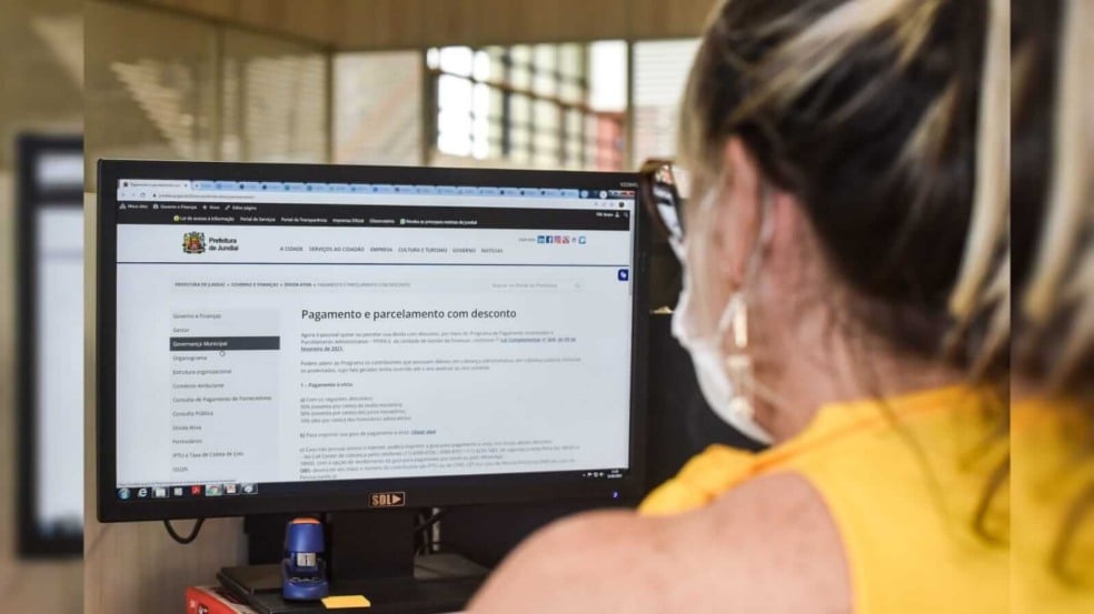 Uma mulher de blusa amarela sentada na frente do computador que está aberto no site da prefeitura de Jundiaí
