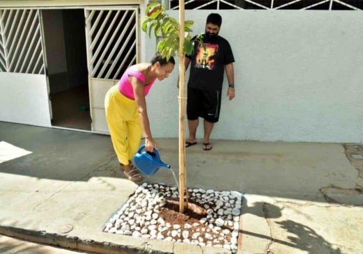 Mulher regando uma árvore recém-plantada em frente a uma casa. Ela está usando uma blusa rosa e calças amarelas, enquanto segura um regador azul. Um homem, que parece observá-la, está ao fundo, vestindo uma camiseta preta com uma estampa colorida e bermuda preta. A área ao redor da base da árvore é coberta com pedras brancas.