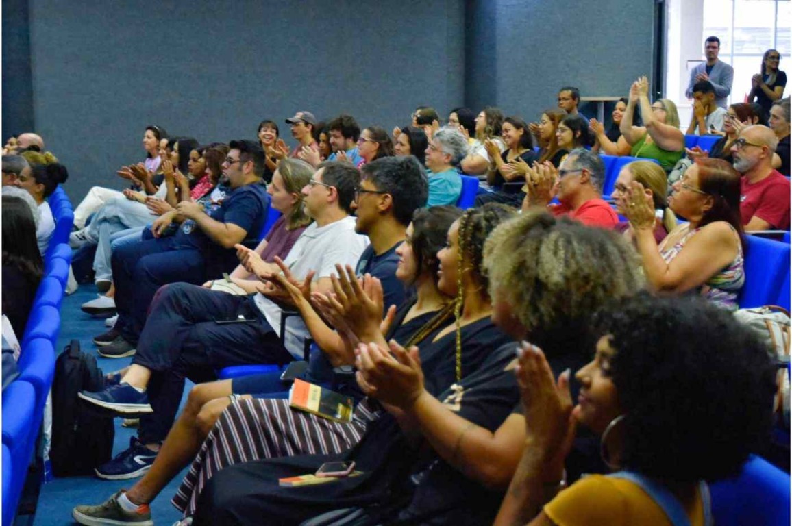 Público acompanha sessão cultural na Biblioteca de Jundiaí, com auditório lotado e pessoas aplaudindo animadas.