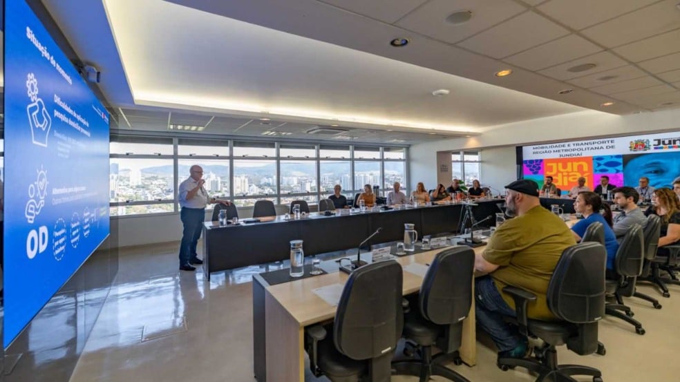 Reunião na Prefeitura de Jundiaí com representantes da Região Metropolitana, onde um palestrante apresenta dados sobre mobilidade em um telão, diante de participantes sentados em mesa de conferência com vista da cidade ao fundo.