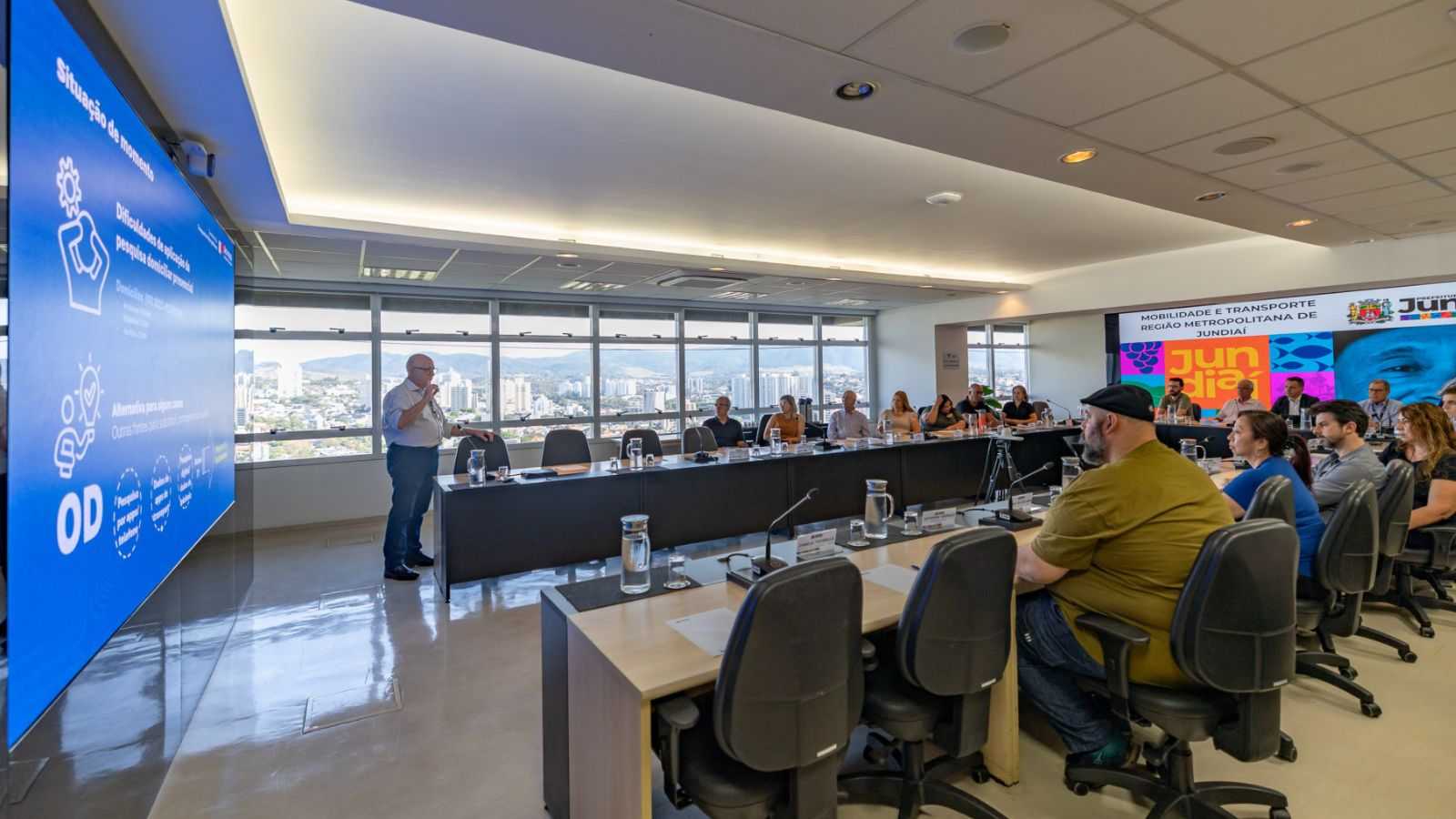 Reunião na Prefeitura de Jundiaí com representantes da Região Metropolitana, onde um palestrante apresenta dados sobre mobilidade em um telão, diante de participantes sentados em mesa de conferência com vista da cidade ao fundo.