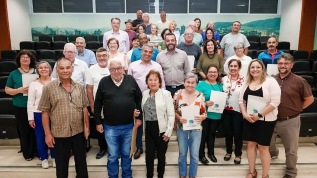 Grupo de moradores de Jundiaí posa sorridente em auditório durante cerimônia de entrega de matrículas de Regularização Fundiária, segurando documentos oficiais.