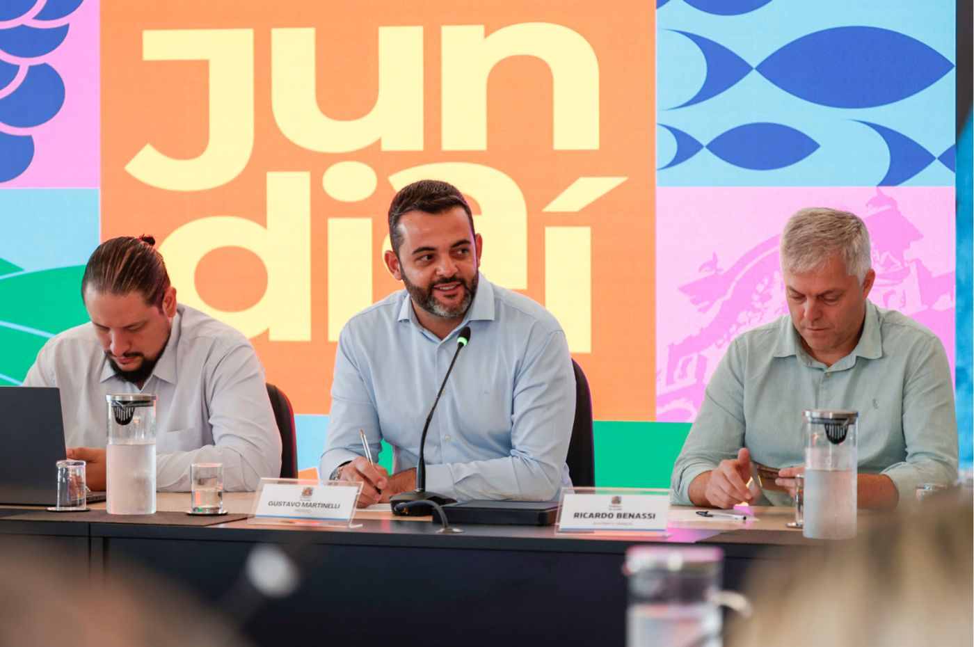 Gustavo Martinelli fala durante reunião da Prefeitura de Jundiaí sobre combate à dengue, com painel colorido ao fundo.