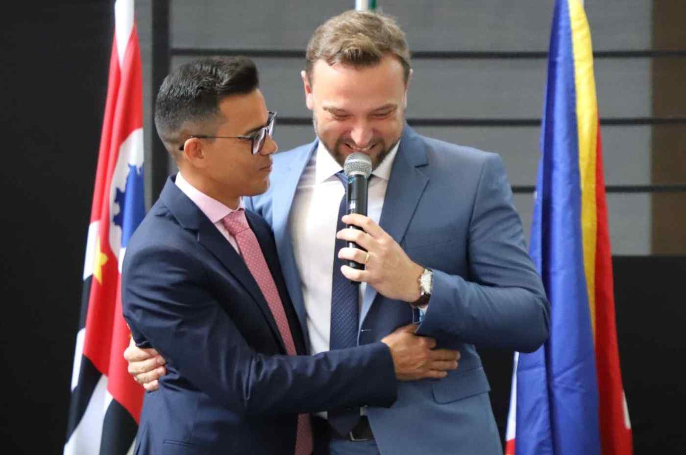 Rogério Cavalin discursa ao microfone enquanto é abraçado por Isaque Messias em cerimônia de posse na Prefeitura de Itupeva.