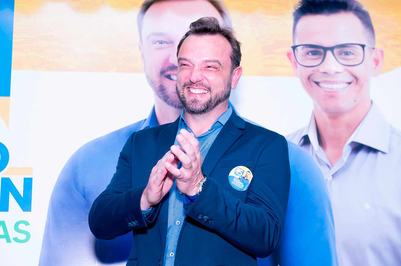 Rogério Cavalin, prefeito de Itupeva, sorrindo e aplaudindo durante evento de campanha em Itupeva, com banner ao fundo destacando sua imagem.
