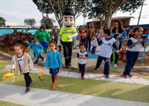 Crianças e adultos participam de uma atividade na Área da Infância em Jundiaí, acompanhadas por mascotes e segurando fitas coloridas.